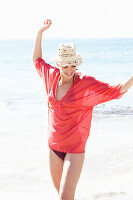 Reife brünette Frau mit rotem Shirt, schwarzem Badenanzug und beigem Hut am Strand