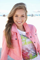 A young blonde woman on a beach wearing a printed t-shirt and a pink denim jacket