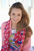 A young blonde woman on a beach wearing a printed t-shirt, a pink denim jacket and a patterned scarf