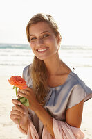 A mature blonde woman on a beach wearing a silver summer dress and holding an orange flower