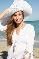 Junge blonde Frau in weißem Sommerkleid, weißem Sommerhut und pinker Halskette am Strand