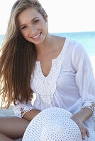 A young blonde woman on a beach wearing a white summer dress and a white summer hat