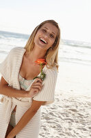 Reife blonde Frau in Dessous und Strickjacke und mit Blume in der Hand am Strand