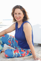 A brunette woman on a beach wearing a purple top and colourful trousers