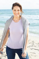 Brunette woman wearing lilac T-shirt, jeans and grey sweater over shoulders on beach