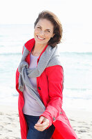 Brunette woman wearing read trench coat with grey sweater over shoulders on beach