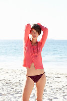 A mature brunette woman on a beach wearing a black bikini and a red shirt