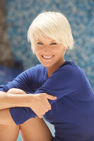 Reife, kurzhaarige blonde Frau im blauen Shirt vor blauem Hintergrund