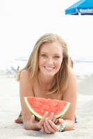 A young blonde woman on a beach wearing a colourful summer dress holding a wedge of melon