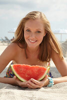 A young blonde woman on a beach wearing a colourful summer dress holding a wedge of melon