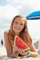 A young blonde woman on a beach wearing a colourful summer dress holding a wedge of melon