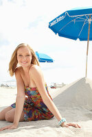 A young blonde woman on a beach wearing a colourful summer dress holding a wedge of melon