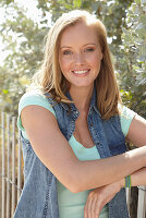 A young blonde woman outside wearing a sleeveless denim jacket and a light-blue top