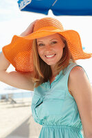 Junge blonde Frau im hellblauen Sommerkleid und orangenem Sommerhut am Strand