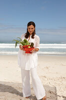 A young brunette woman on a beach wearing a white summer dress and holding a bowl of vegetables