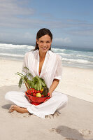 A young brunette woman on a beach wearing a white summer dress and holding a bowl of vegetables