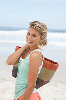 Reife blonde Frau im weißen Top, türkisfarbener Weste und Bastkorb am Strand