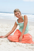 Reife blonde Frau im weißen Top, türkisfarbener Weste und lachsfarbenem Rock am Strand
