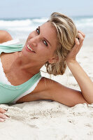 Reife blonde Frau im weißen Top und türkisfarbener Weste am Strand
