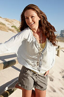 A brunette woman on a sandy beach wearing a light blouse and shorts