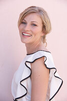 A blonde woman standing against a pink background wearing a light blouse with a black trim