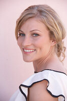 A blonde woman standing against a pink background wearing a skirt and a light blouse with a black trim