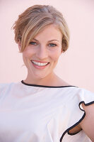 A blonde woman standing against a pink background wearing a skirt and a light blouse with a black trim