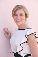 A blonde woman standing against a pink background wearing a skirt and a light blouse with a black trim