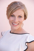 A blonde woman standing against a pink background wearing a skirt and a light blouse with a black trim