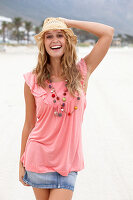 Junge blonde Frau im rosa Top, kurzem Jeansrock und beigem Hut am Strand