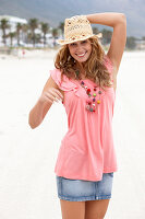 A young blonde woman on a beach wearing a pink top, a short denim skirt and a beige hat