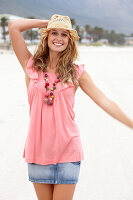 Junge blonde Frau im rosa Top, kurzem Jeansrock und beigem Hut am Strand