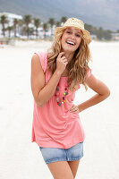 A young blonde woman on a beach wearing a pink top, a short denim skirt and a beige hat