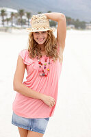 A young blonde woman on a beach wearing a pink top, a short denim skirt and a beige hat