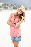 Junge blonde Frau im rosa Top, kurzem Jeansrock und beigem Hut am Strand