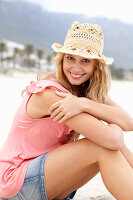 Junge blonde Frau im rosa Top, kurzem Jeansrock und beigem Hut am Strand