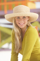 A young blonde woman on a beach wearing an olive top and a beige summer hat