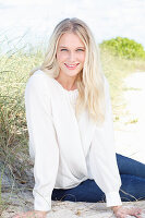 A young blonde woman sitting in the sand wearing a white blouse and jeans