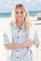 Junge blonde Frau in gestreiftem Hemd und mit weißem Pulli am Strand
