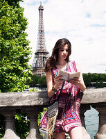 A young brunette woman wearing a summer dress looking at a guide book on a bridge near the Eiffel Tower