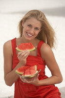 A young blonde woman wearing a red summer dress holding slices of watermelon