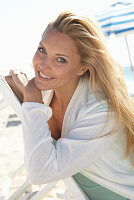 A blonde woman on a beach wearing a turquoise top and a white jacket