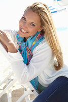 A blonde woman on a beach wearing a colourful scarf and a white jacket