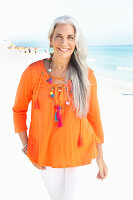 A mature woman with white hair on a beach wearing an orange tunic and white summer trousers