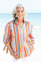 A mature woman with white hair on a beach wearing a striped shirt and white summer trousers