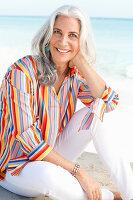 A mature woman with white hair on a beach wearing a striped shirt and white summer trousers