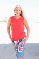 A mature woman with white hair on a beach wearing a red top and a pair of floral-patterned summer trousers