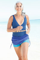 A mature woman with white hair on a beach wearing a blue bathing suit and turquoise shorts