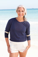 A mature woman with white hair on a beach wearing a blue top and white shorts