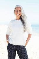 A mature woman with white hair on a beach wearing a white jumper and polka dot trousers